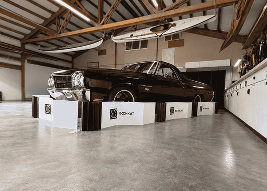 A black vintage car displayed inside a Box-Kat mouse barrier with exposed wooden ceiling beams and a concrete floor.